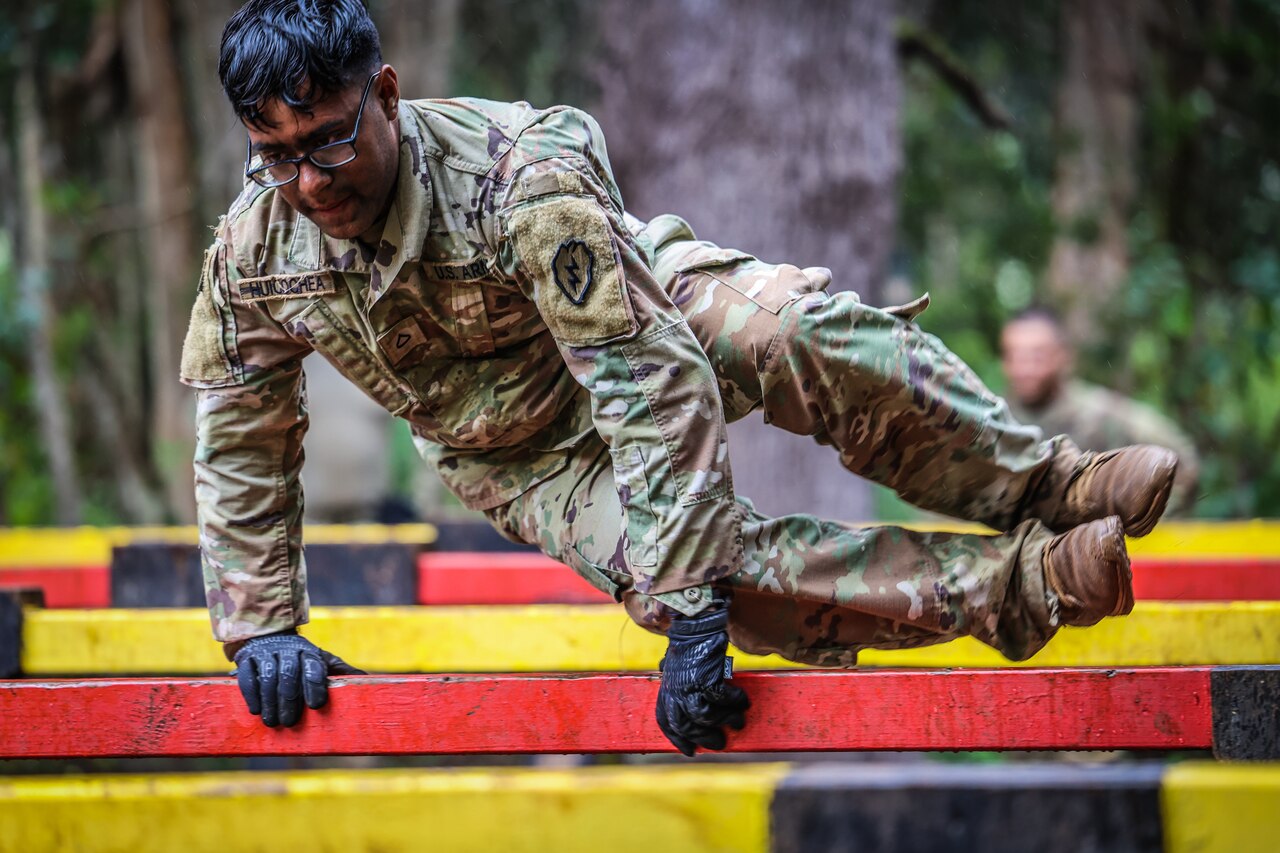 A soldier wearing gloves jumps over a red wooden bar.