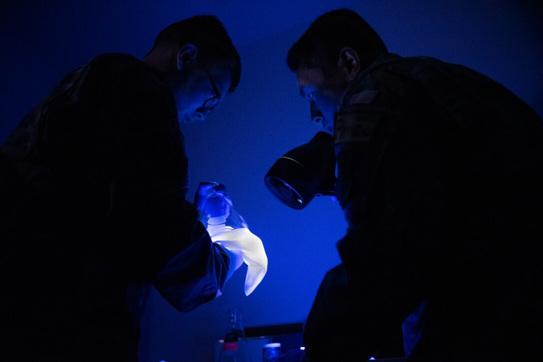 Two men work in a darkened setting, one holds a large flashlight.