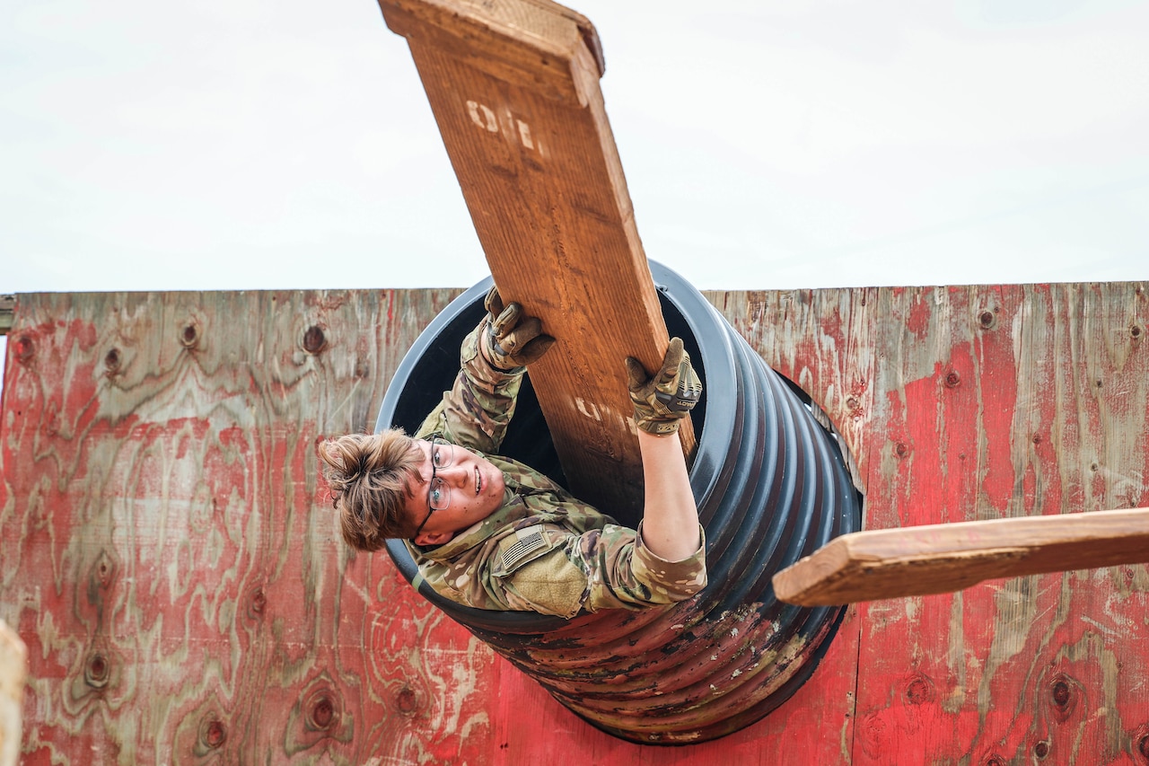 A soldier emerges head first from a raised tube and holds a wooden plank.