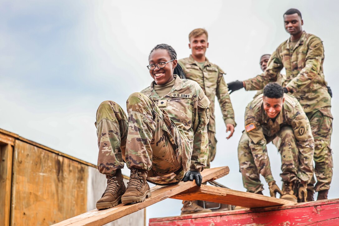 A soldier smiles while sitting on a raised wood plank as others stand behind her.
