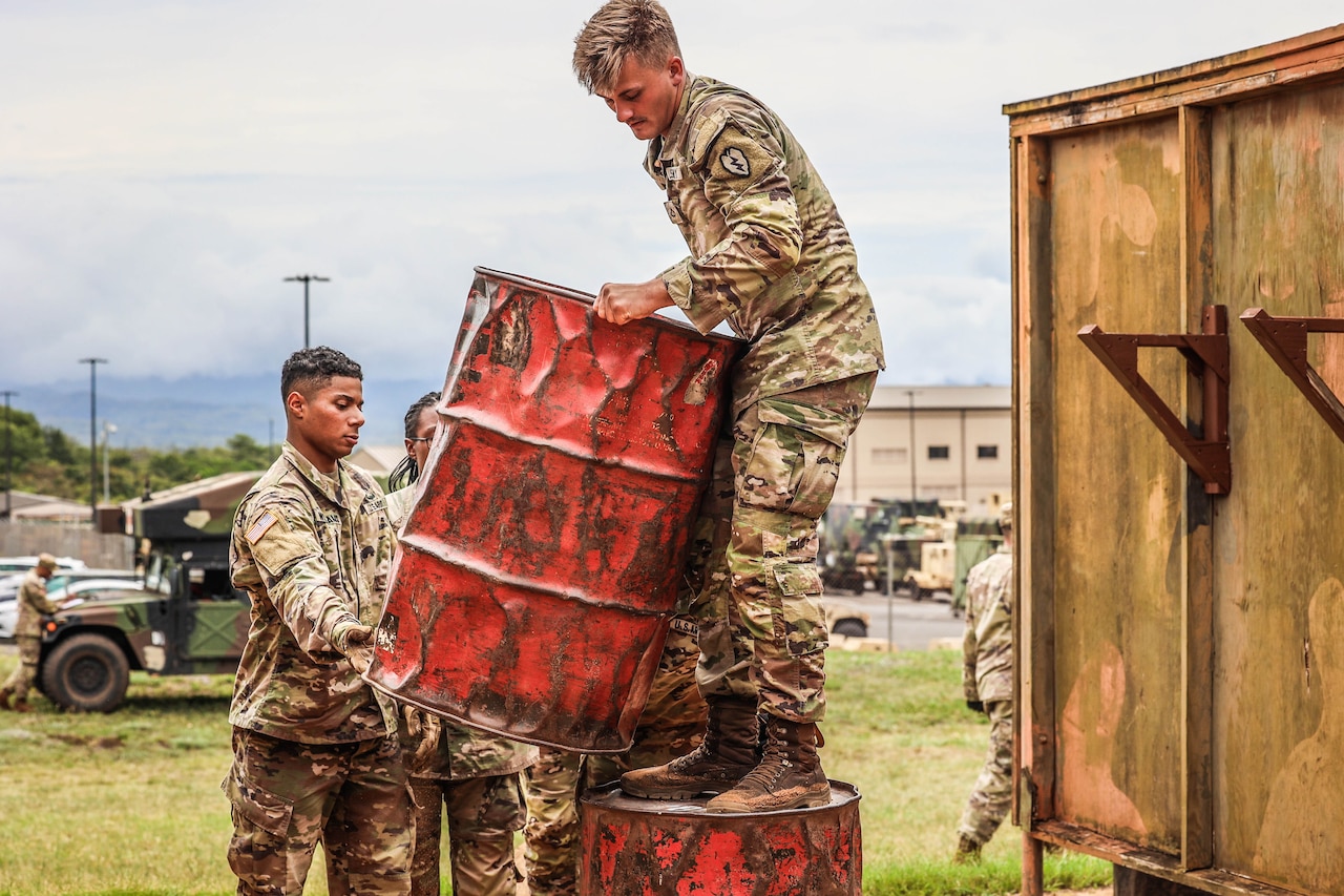 A soldier stands on an orange metal drum as fellow soldiers help him lift another drum.