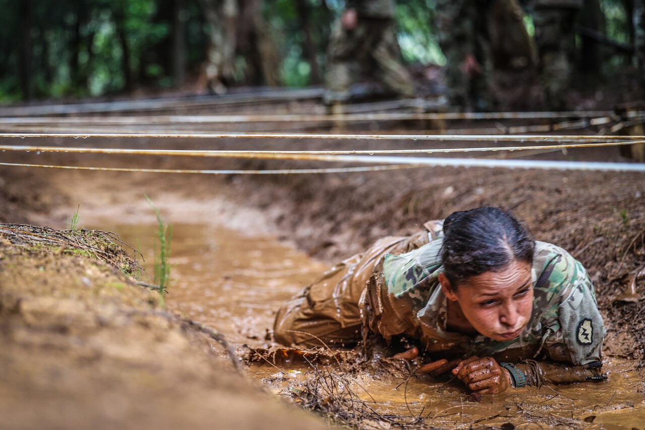 A soldier crawls in mud under wires.