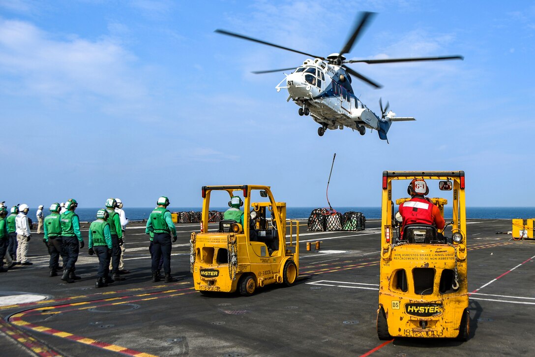 A helicopter drops a pallet onto the deck of a ship while a group of sailors wait.