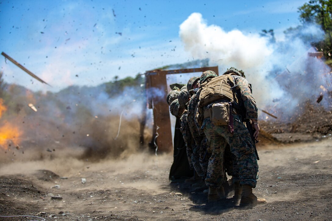 A line of Marines crouch behind each other as a small explosion occurs.