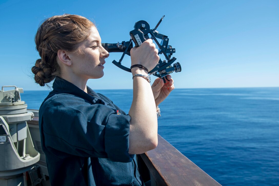 A sailors holds up a sextant on the deck of a ship.