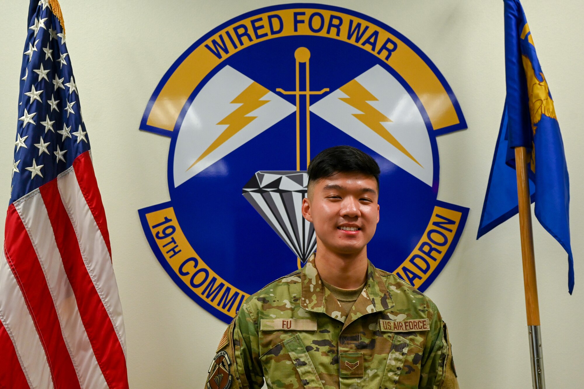 An Airman stands in front of the 19th Communications Squadron emblem.