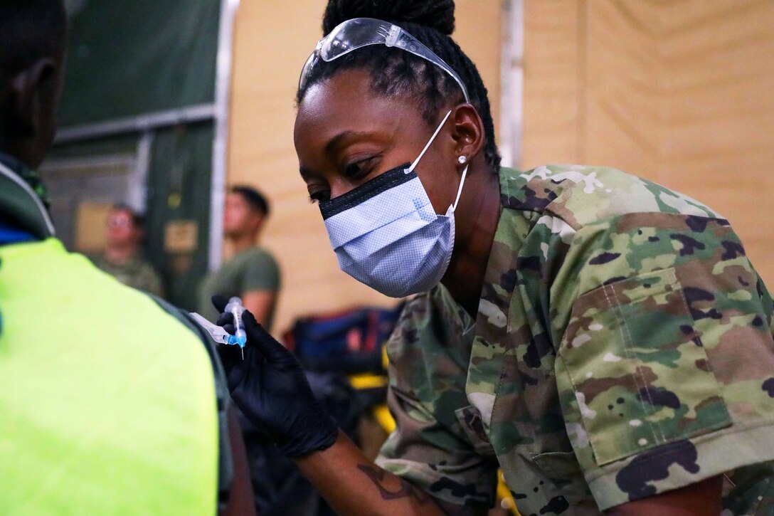 An airman wearing a face mask and gloves leans over to give an injection to a man who's seated next to her.