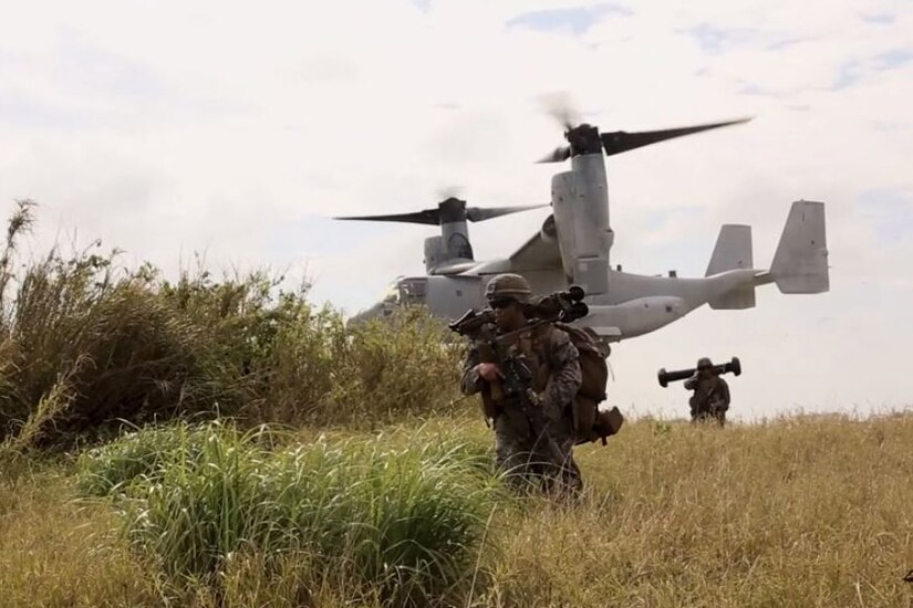 Marines walk through a field as a large aircraft takes off behind them.