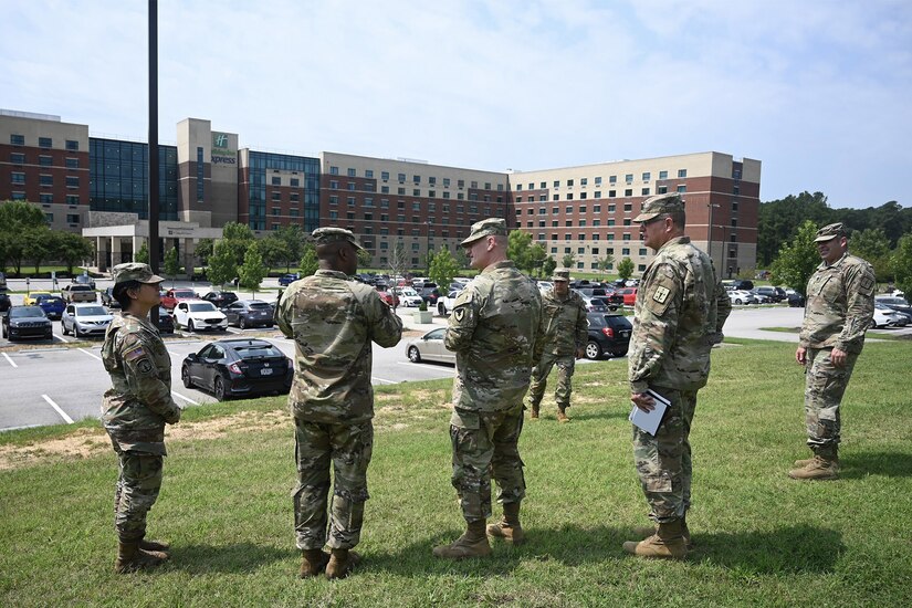 Six soldiers stand on a grassy area near a parking lot in front of a hotel.