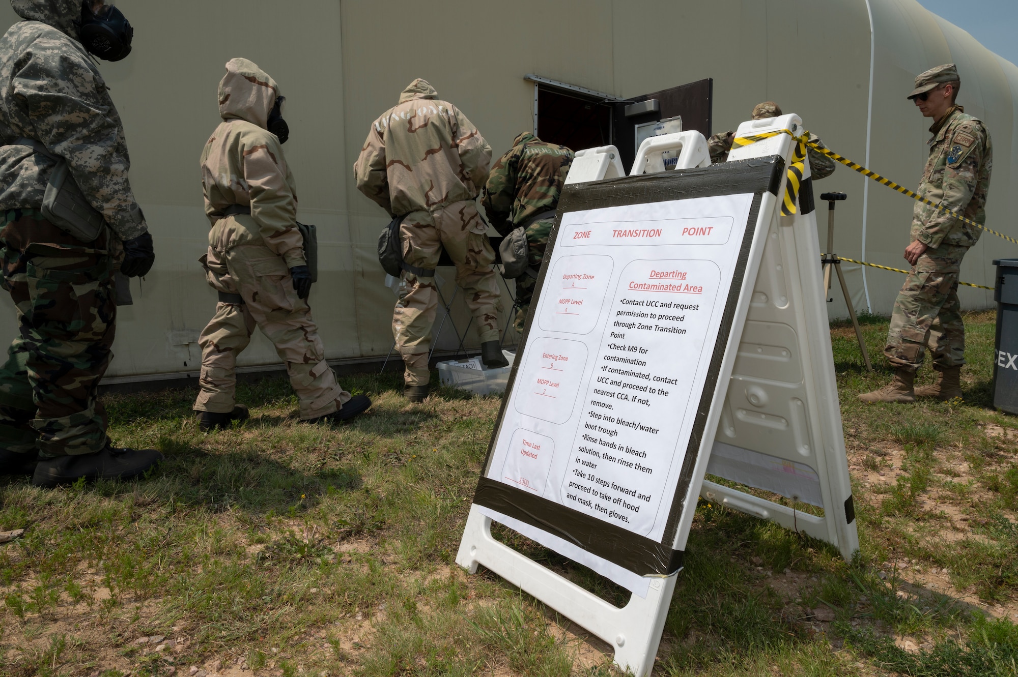 People in chemical gear line up outside a building near a sign with instructions written on it.