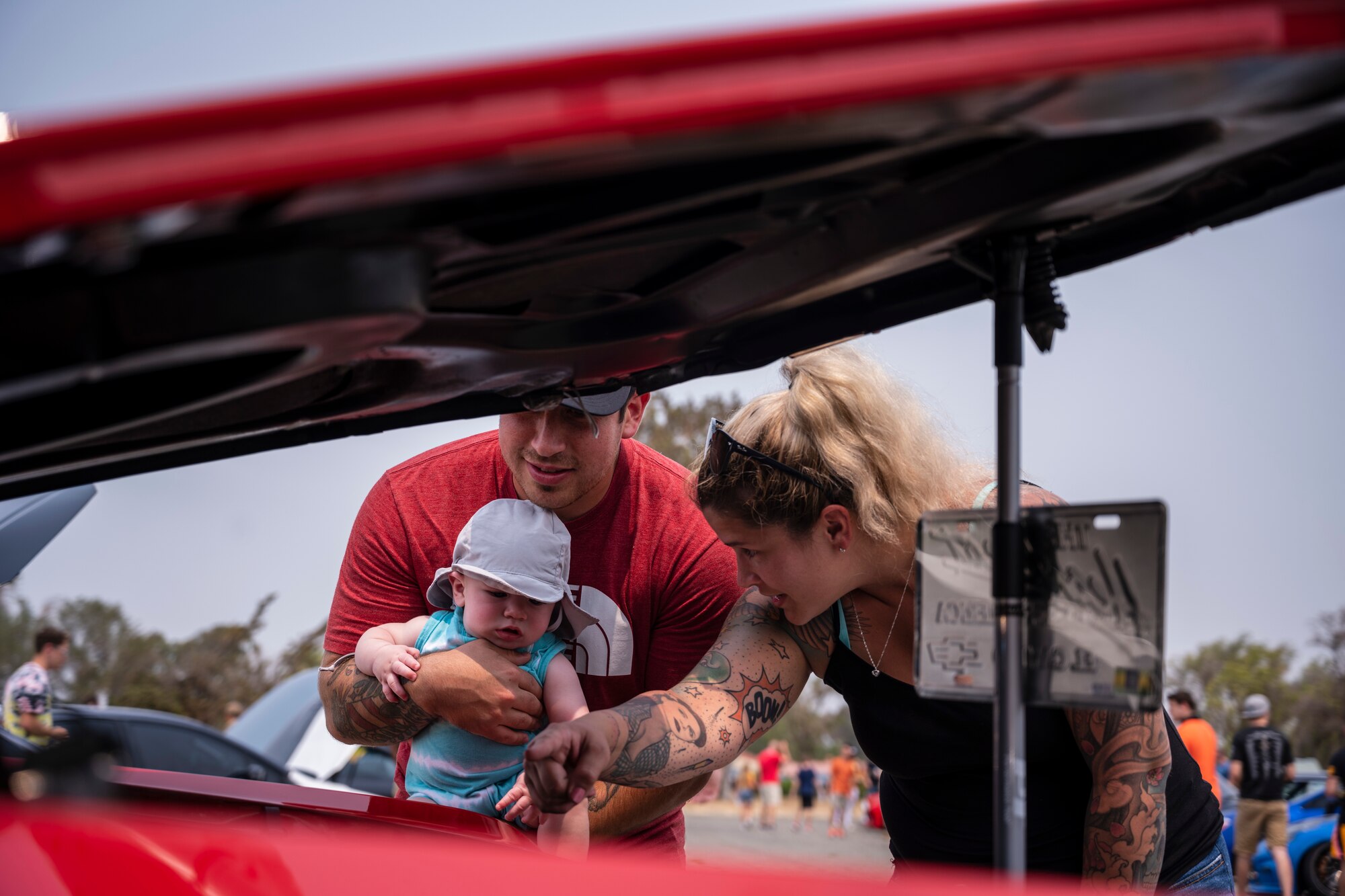 A young family holds a child near a car engine.