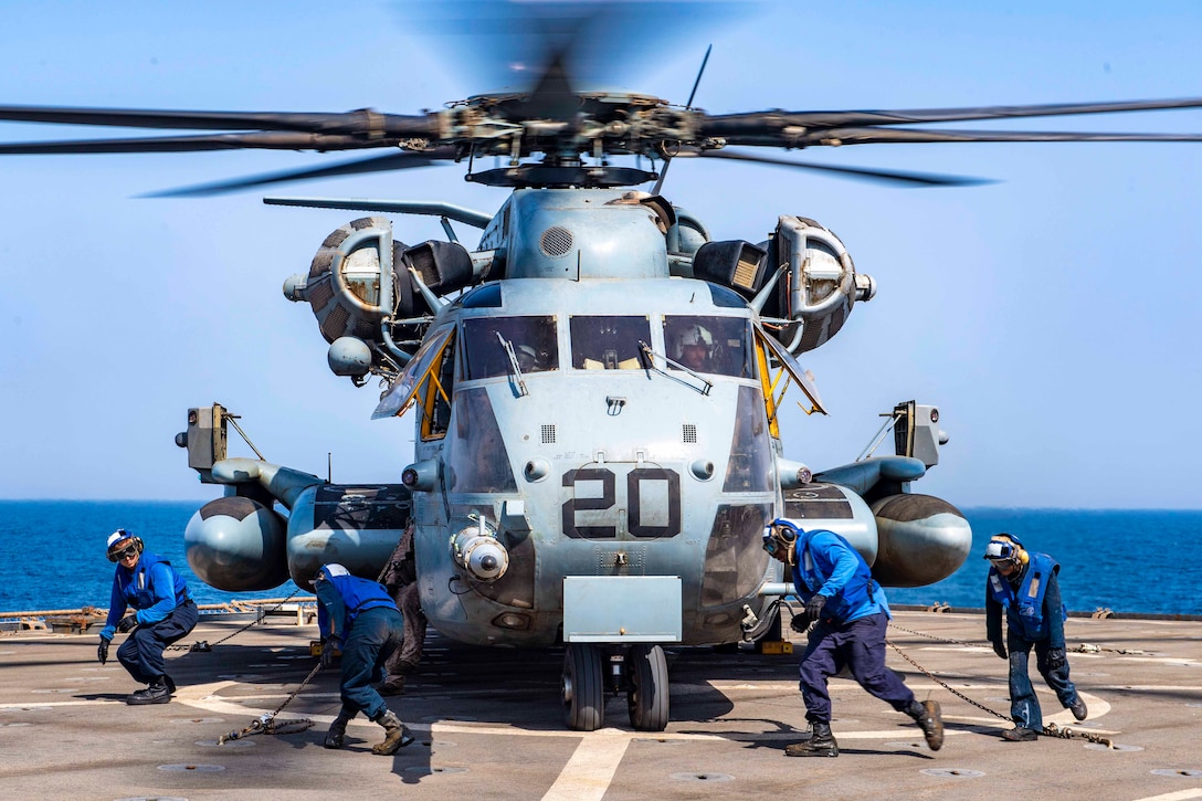 Four sailors run around a helicopter on the deck of a ship.