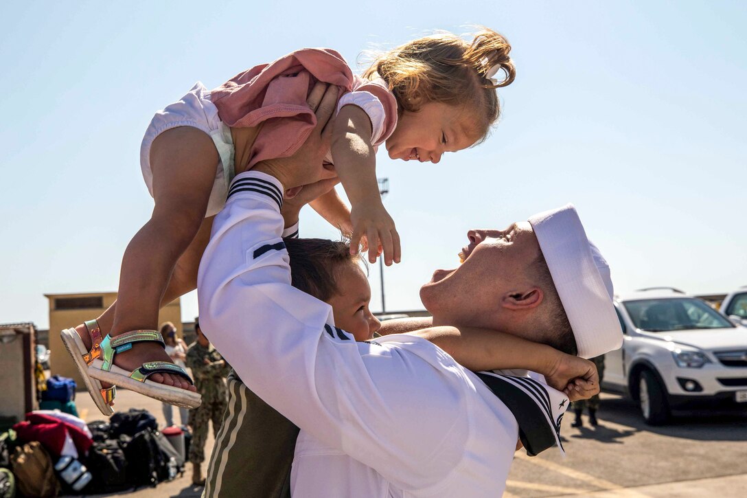 A sailor holds up a toddler while another child hugs him.