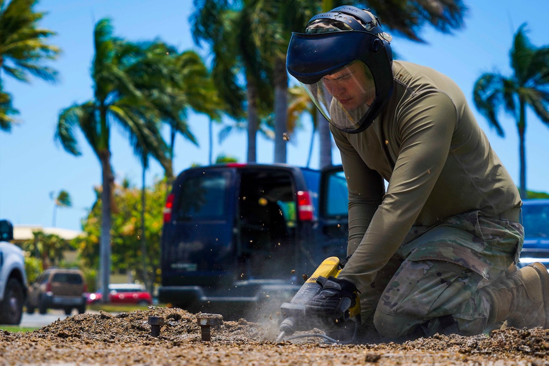 An airman wearing a face guard uses a tool to break up concrete.
