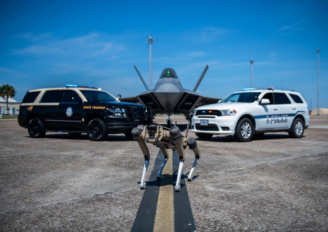 Two cars, a quad-legged unmanned ground vehicle and an airplane are posed for a photo op