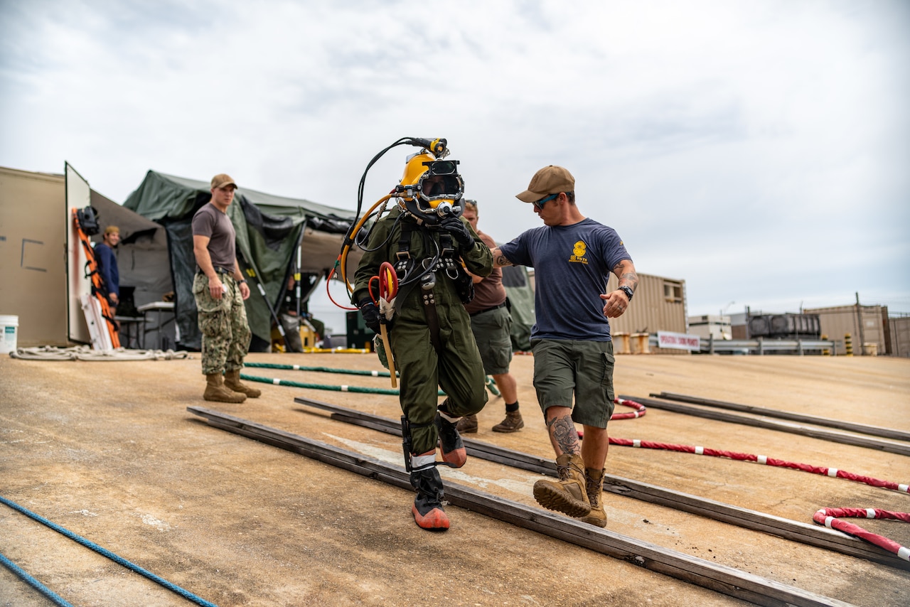 A sailor in dive gear walks next to two others.