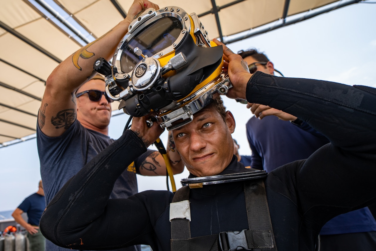 Two people help a sailor put on a dive helmet.
