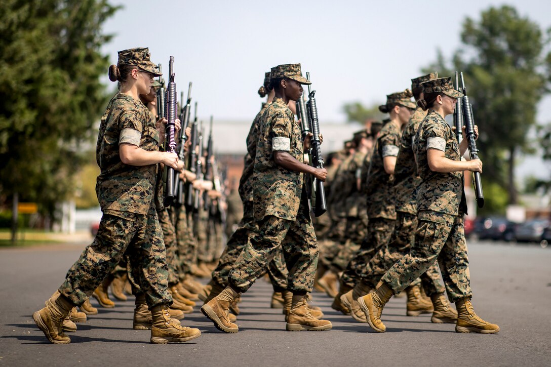 Marines carrying weapons walk on pavement in three rows.