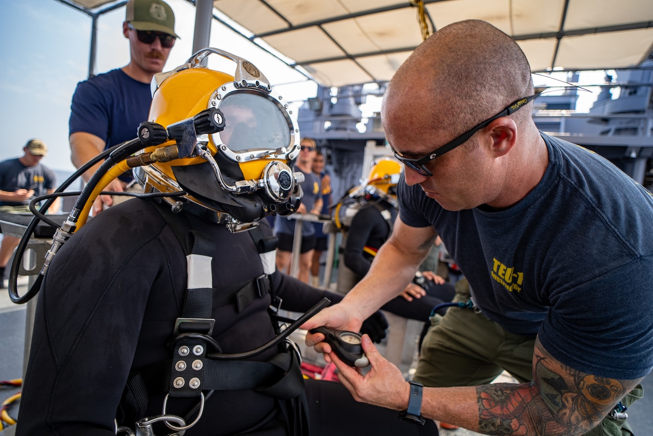 A sailor in dive gear sits as another sailor inspects them.