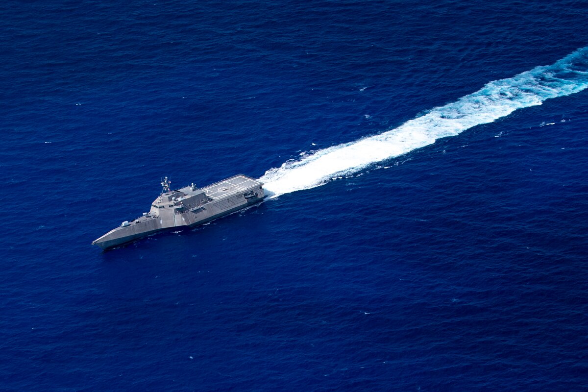 A littoral combat ship, seen from above, creates a white wake while traveling in vivid blue water.