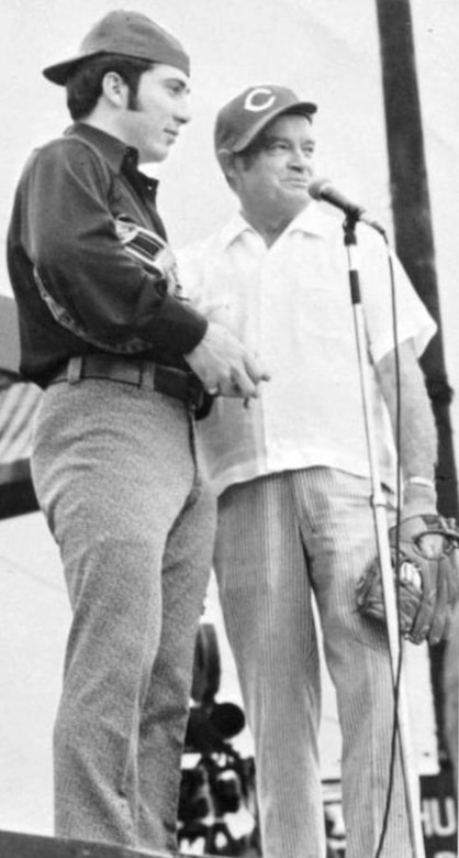 Two men wearing baseball caps stand next to each other; one appears to speak into a microphone.