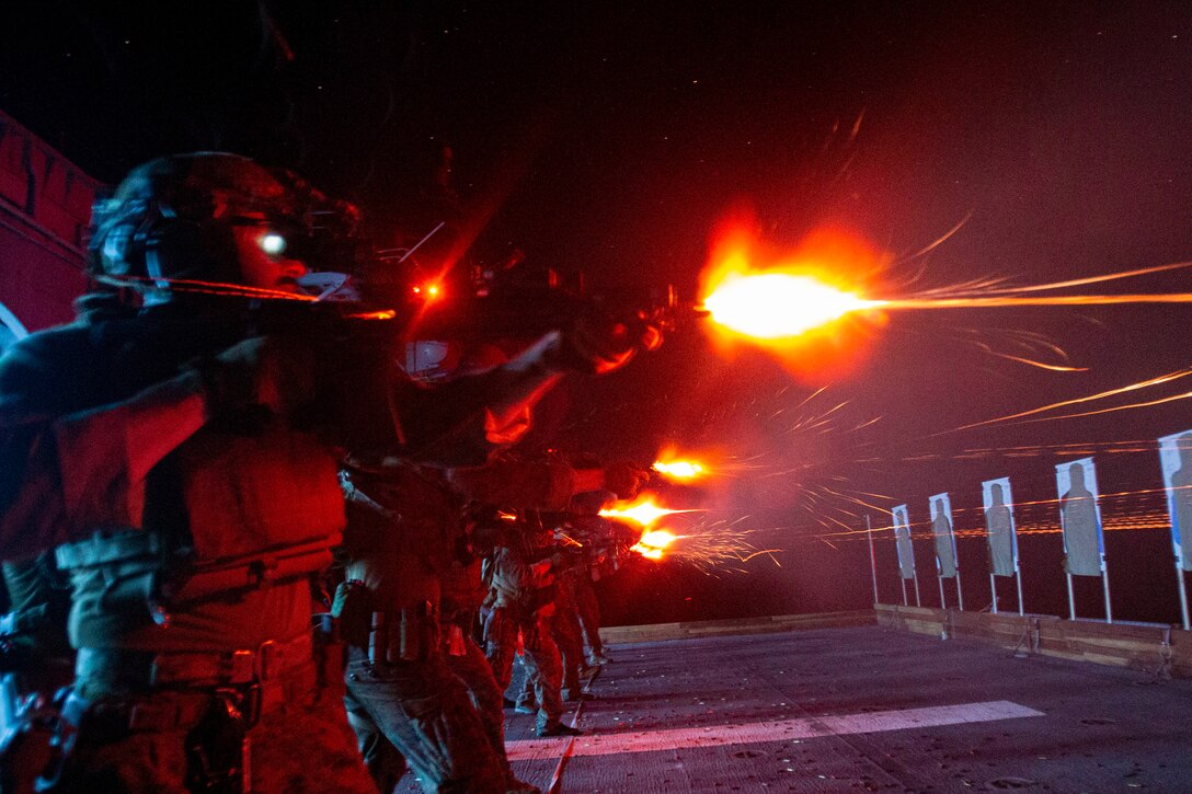 Marines fire at targets at night on a ship's deck, creating red bursts of light.