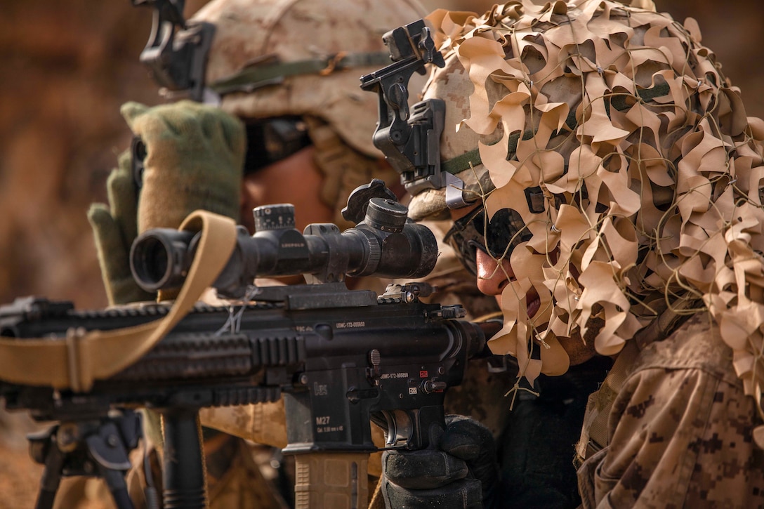 A Marine in camo looks through the scope of a weapon next to another using binoculars.