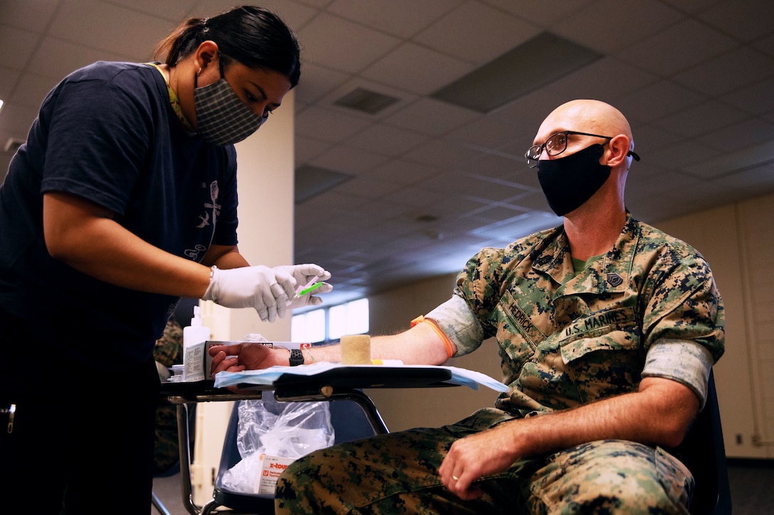 A Marine seated and wearing a face mask donates blood while a woman wearing a face mask and gloves bends over holding a syringe.