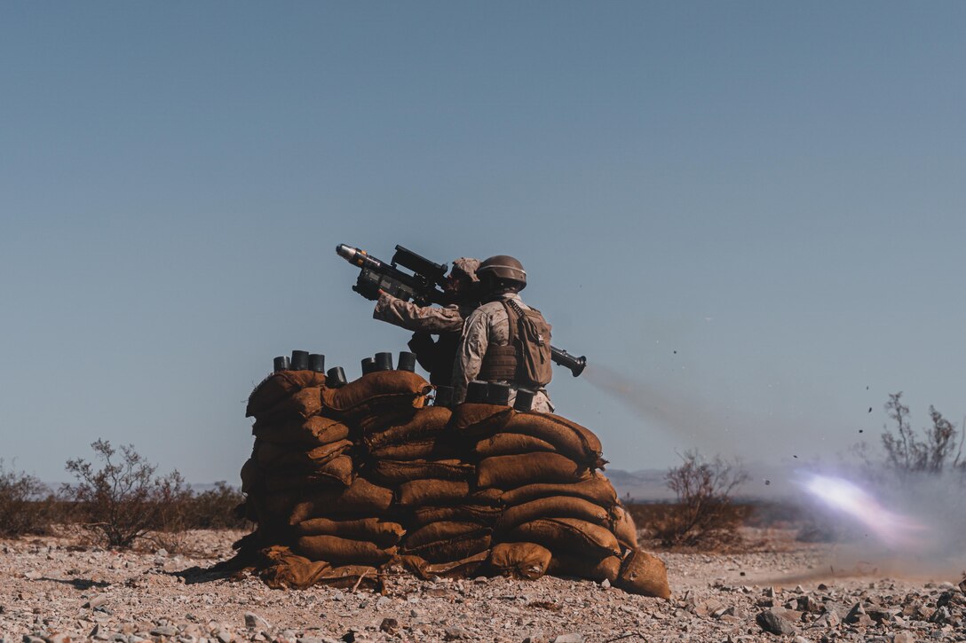 A Marine stands behind a pile of sandbags and fire a missile while another Marine watches.