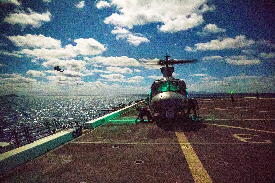 A helicopter sits on the deck of a military ship in the ocean while another flies above.