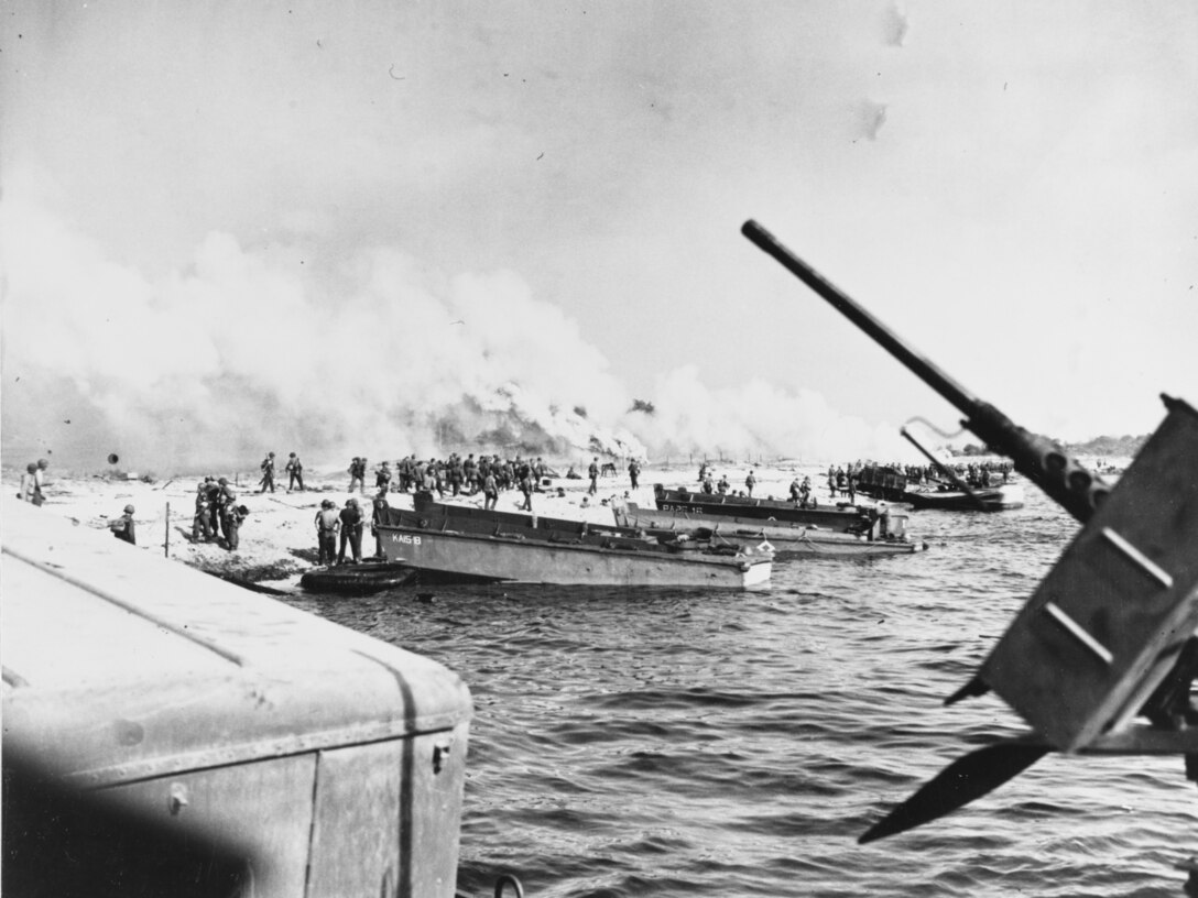 Several small boats pull up to a shoreline. Groups of men are on the beach as plumes of smoke rise nearby.