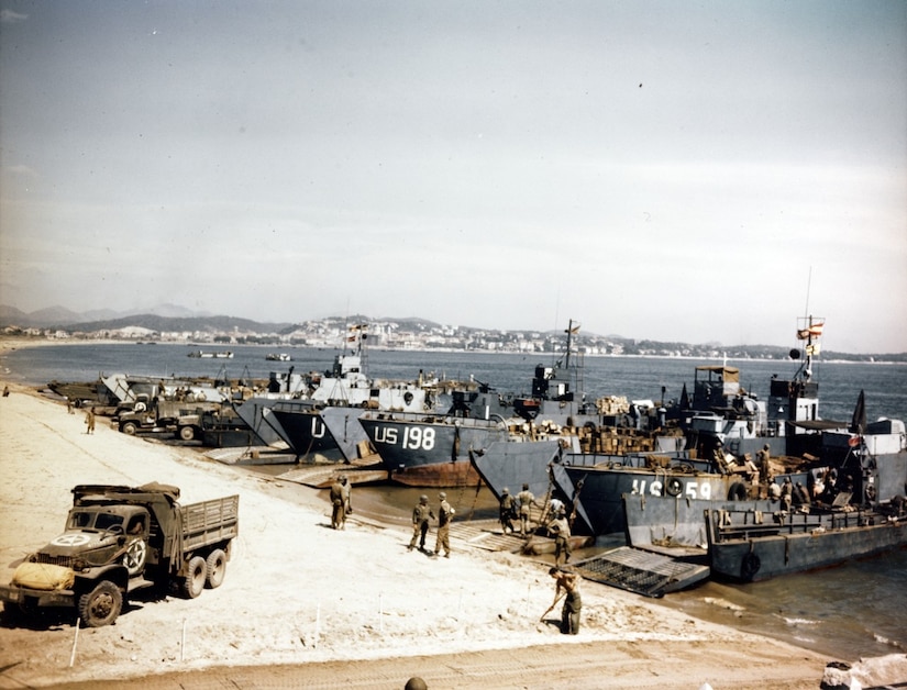 Several landing craft are on a shoreline; a vehicle sits on the beach nearby. Dozens of men work around the vehicles.