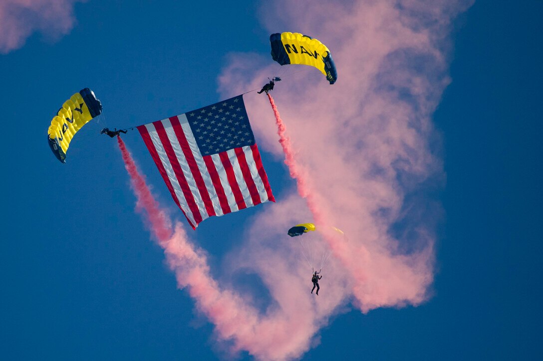 Two sailors display an American flag while freefalling with parachutes as another sailor descends with a parachute behind.