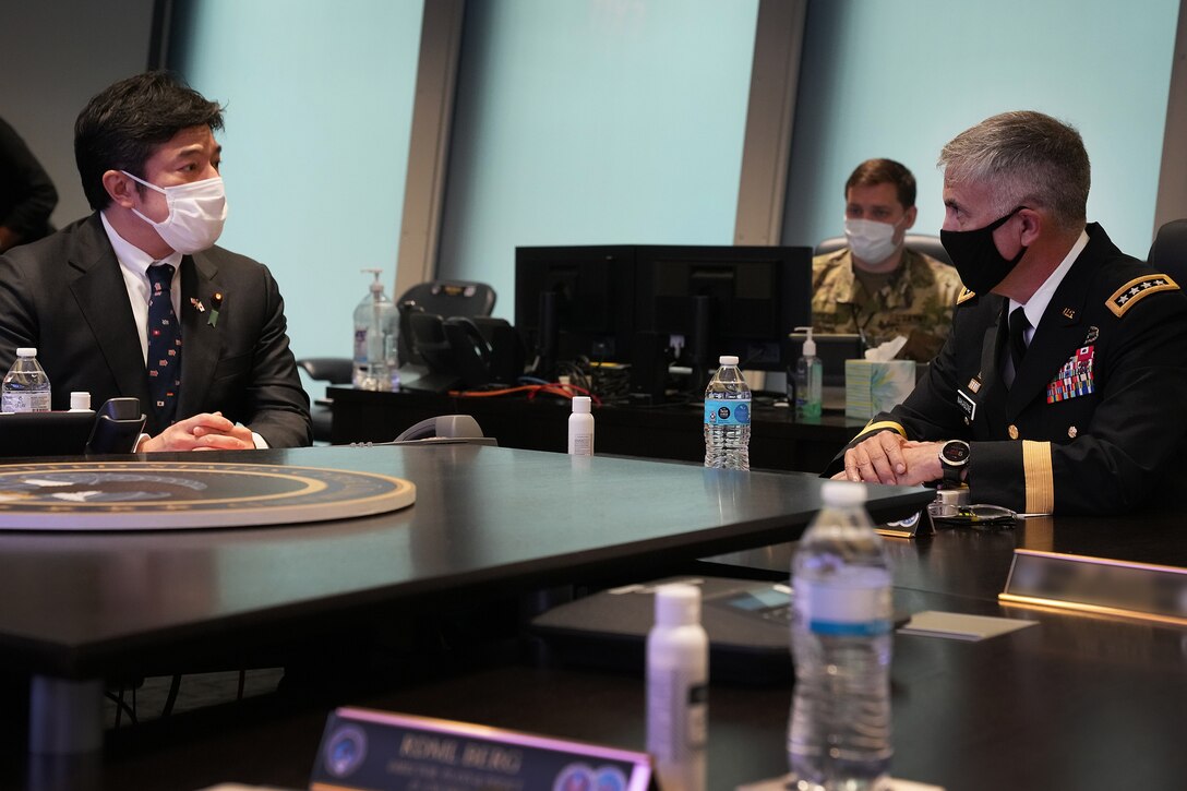 A man in a military uniform and a man in a suit speak while sitting at a conference table.