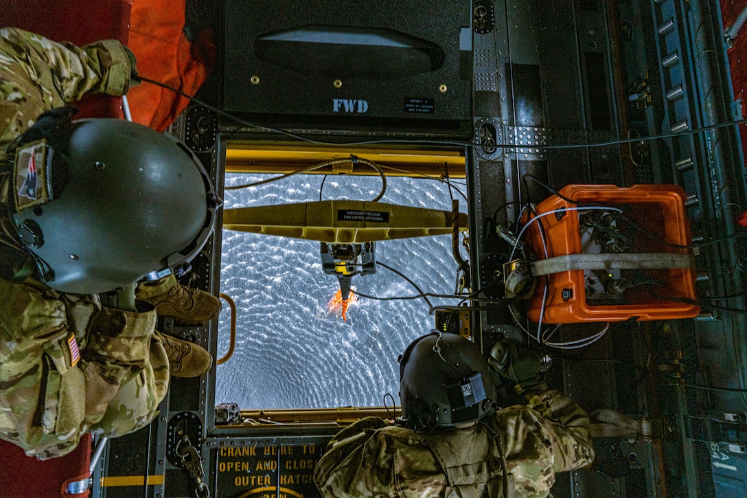 Two soldiers look out of a helicopter hatch at a dangling water bucket and blue water below.