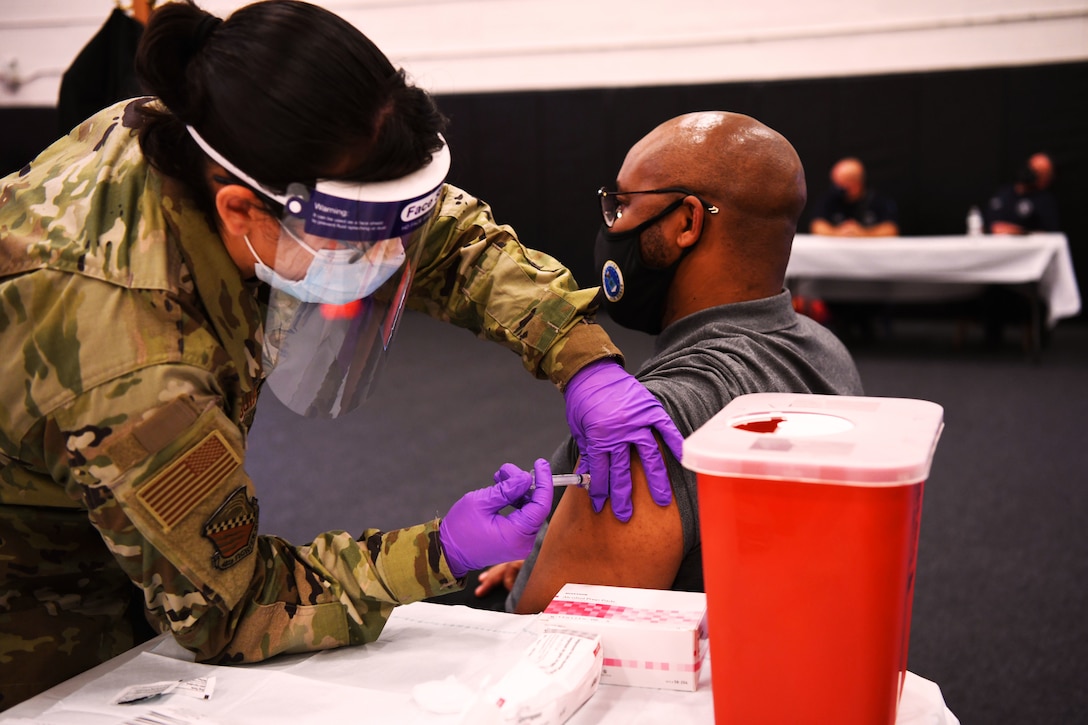 A female service member, who's wearing personal protective equipment, holds a syringe as she leans over to give an injection to a man seated at the end of a table and wearing a mask.