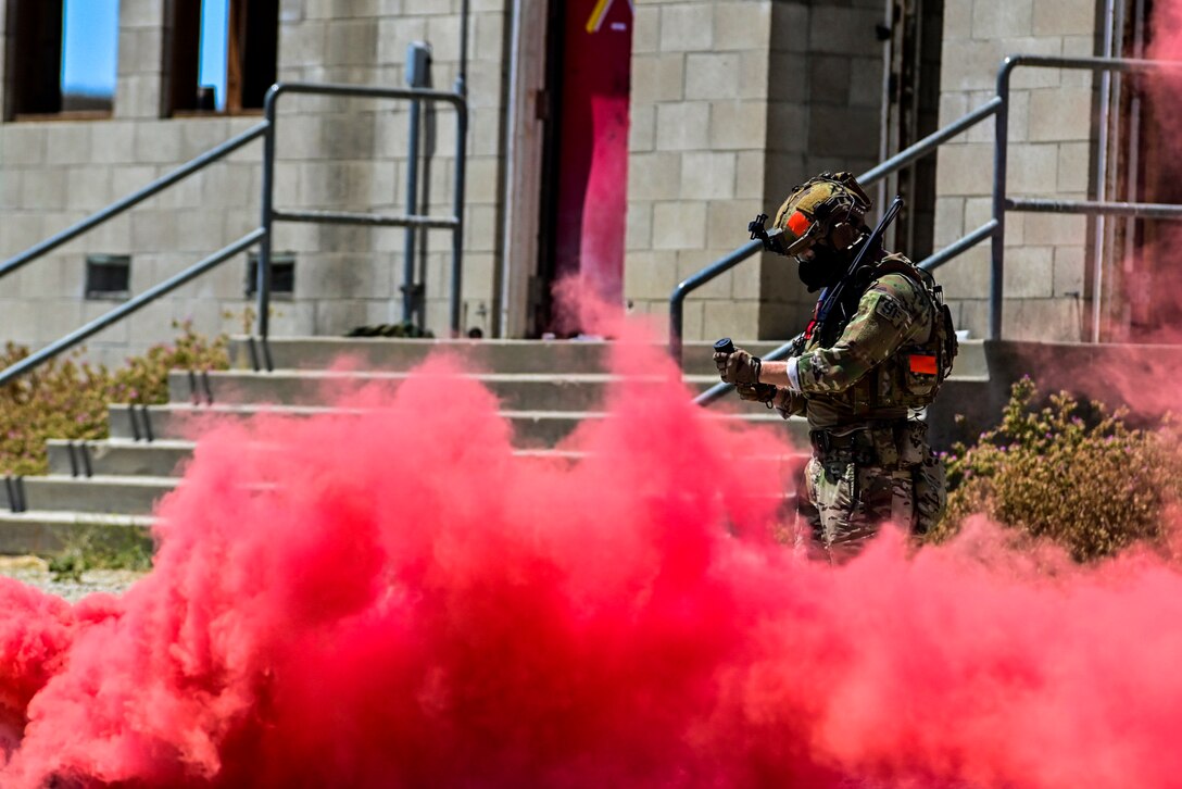 An airman holds a simulated grenade outside a building surrounded by red smoke.