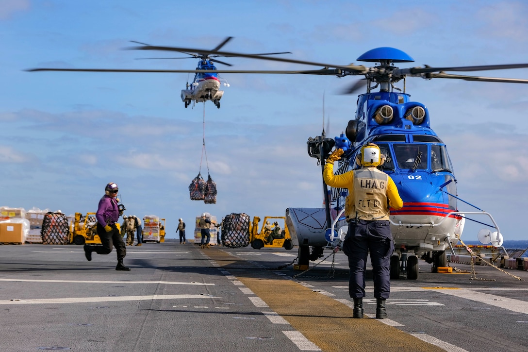 A helicopter carrying cargo hovers over a ship’s deck while a sailor stands next to a parked helicopter.