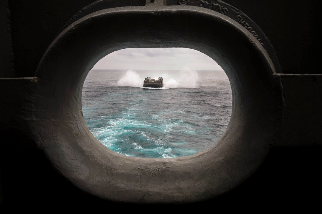 Marines and sailors ride in a small boat behind a ship as seen through the ship’s porthole.