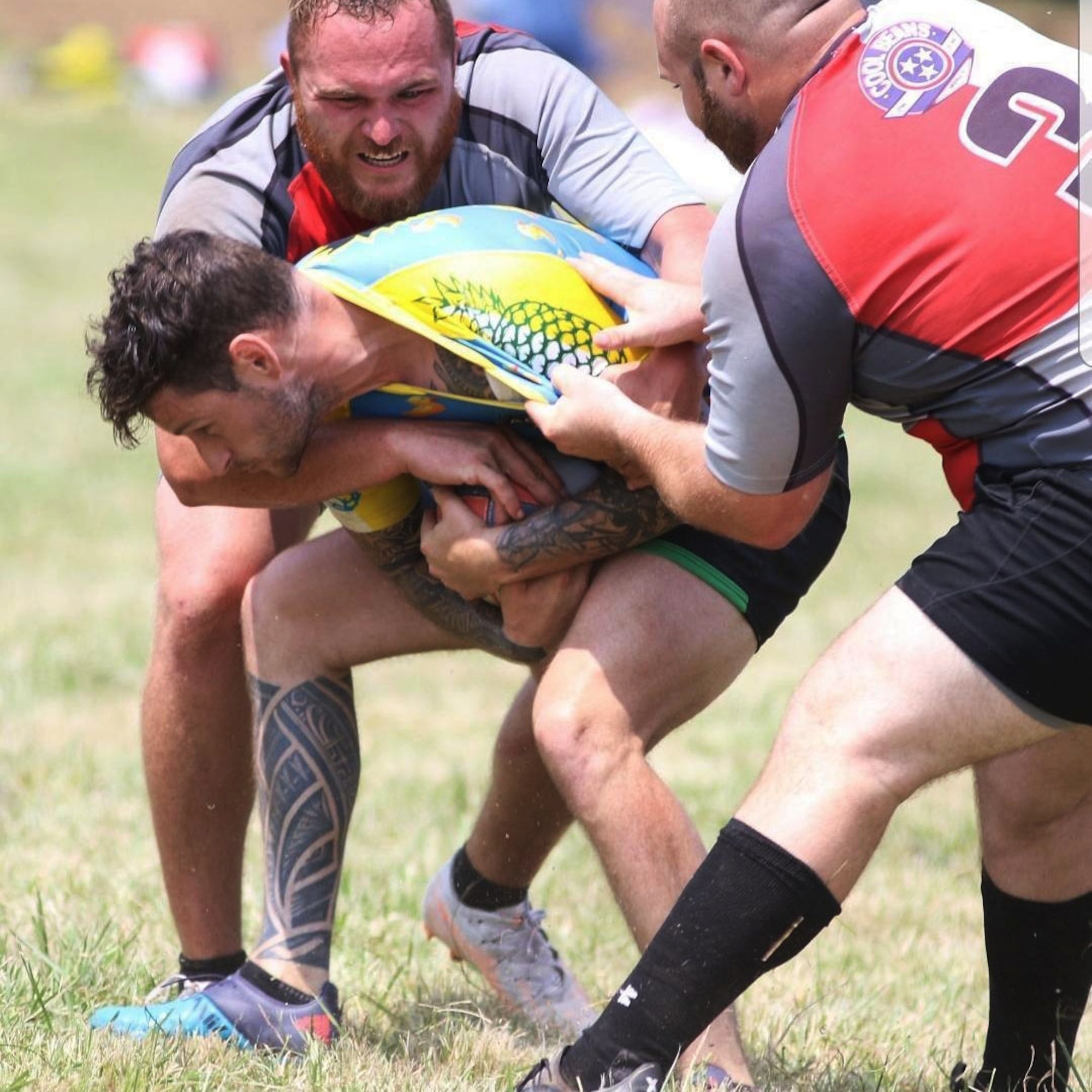 Two men try to take a rugby ball from third man on a grass field.