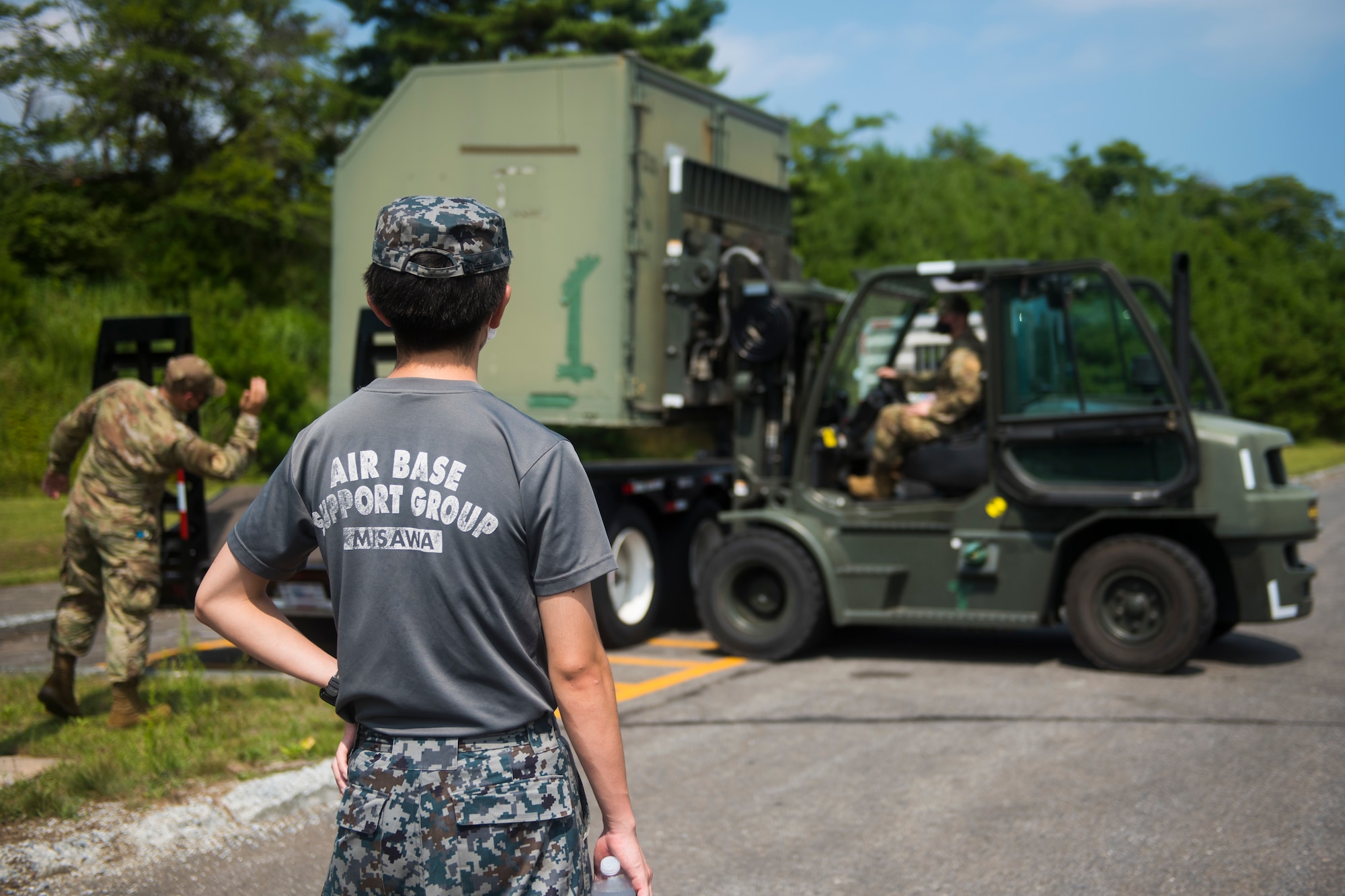 JASDF member watches as USAF members operate a forklift to place cargo on a truck.