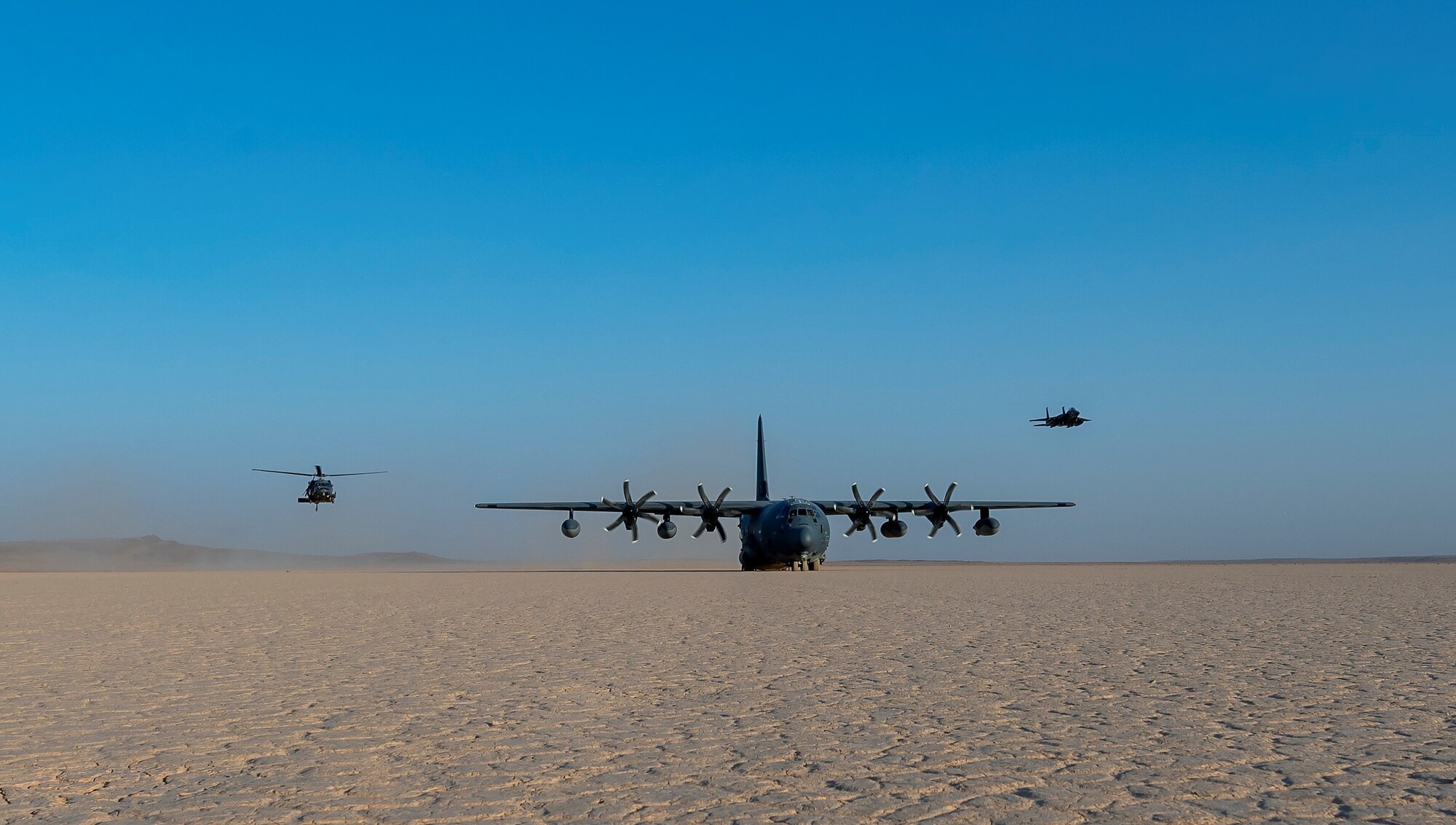 A U.S. Air Force HH-60G Pave Hawk helicopter and an F-15E Strike Eagle fly over a parked HC-130J Combat King II during an exercise July 28, 2021, at an undisclosed location somewhere in Southwest Asia.  The HC-130J provides recovery assistance as well as refueling capabilities at austere locations. (U.S. Air Force photo by Senior Airman Karla Parra)