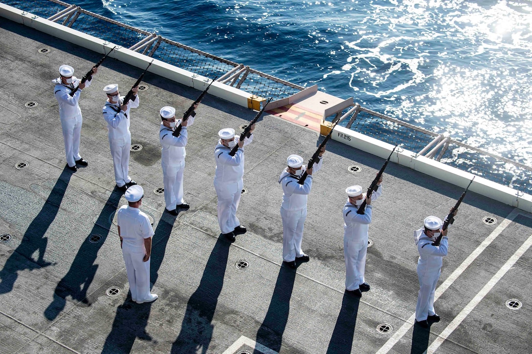 Sailors stand in formation aboard a ship pointing their weapons up.