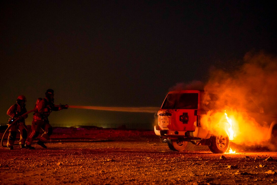 Two firefighters use a hose to spray water on a car fire at night.