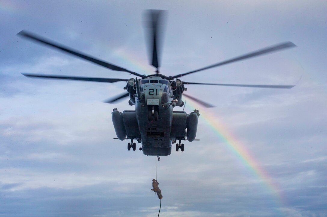 A Marine fast-rope from a helicopter; a rainbow can be seen in the background.