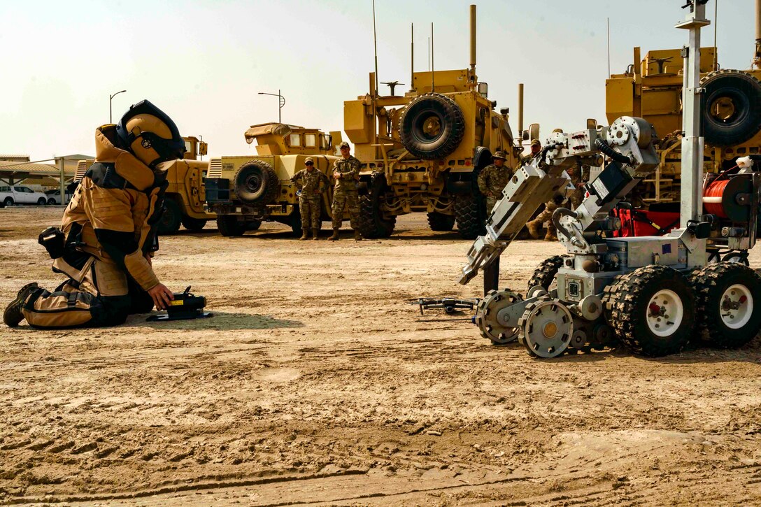 An airman wearing a bomb suit kneels next to a robot.
