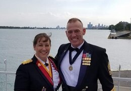 Two Soldiers stand next to each other smiling in their dress mess uniforms with a lake behind them.