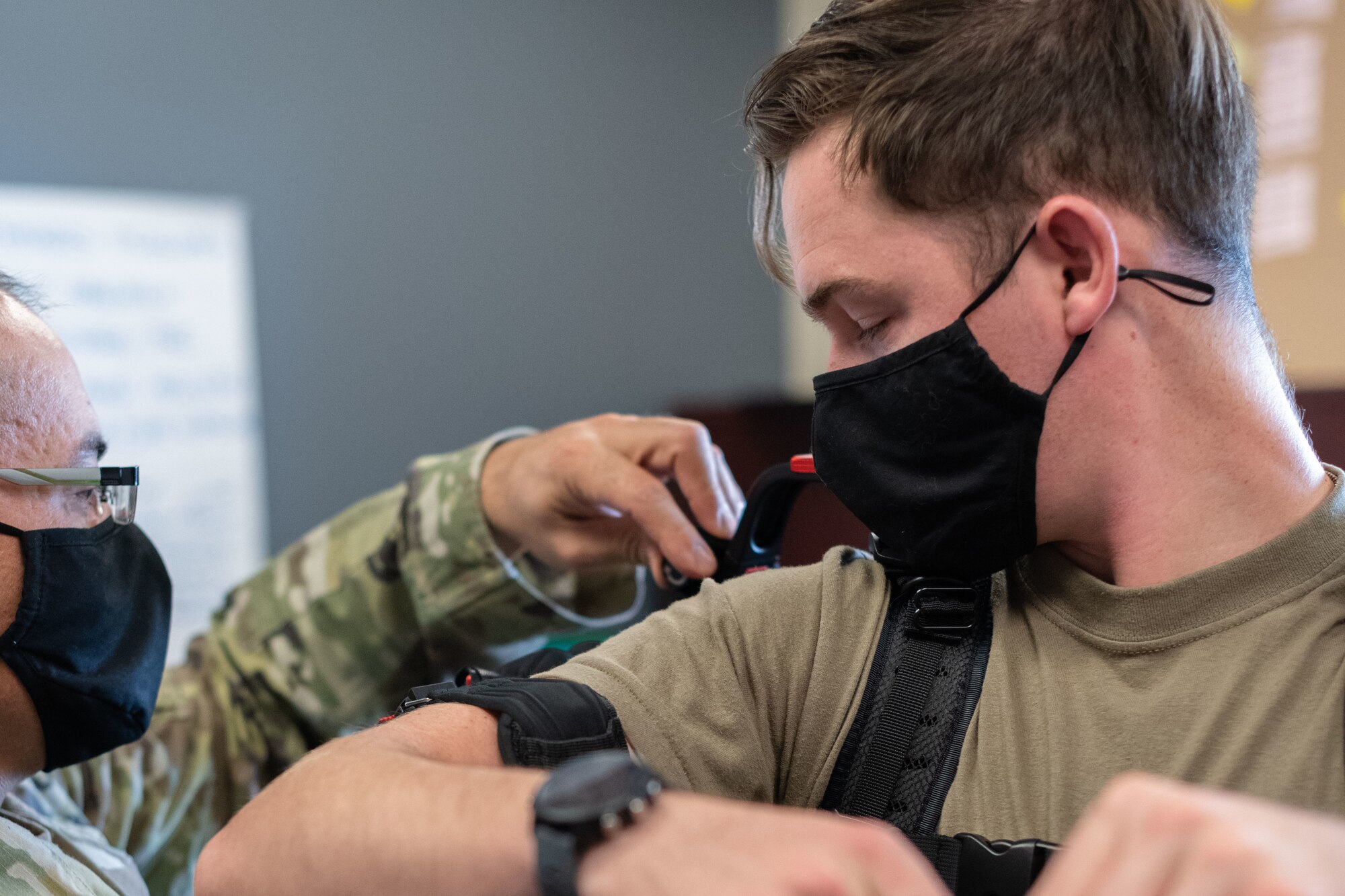An Airman looks on as another Airman straps a suite to his shoulder and arms.
