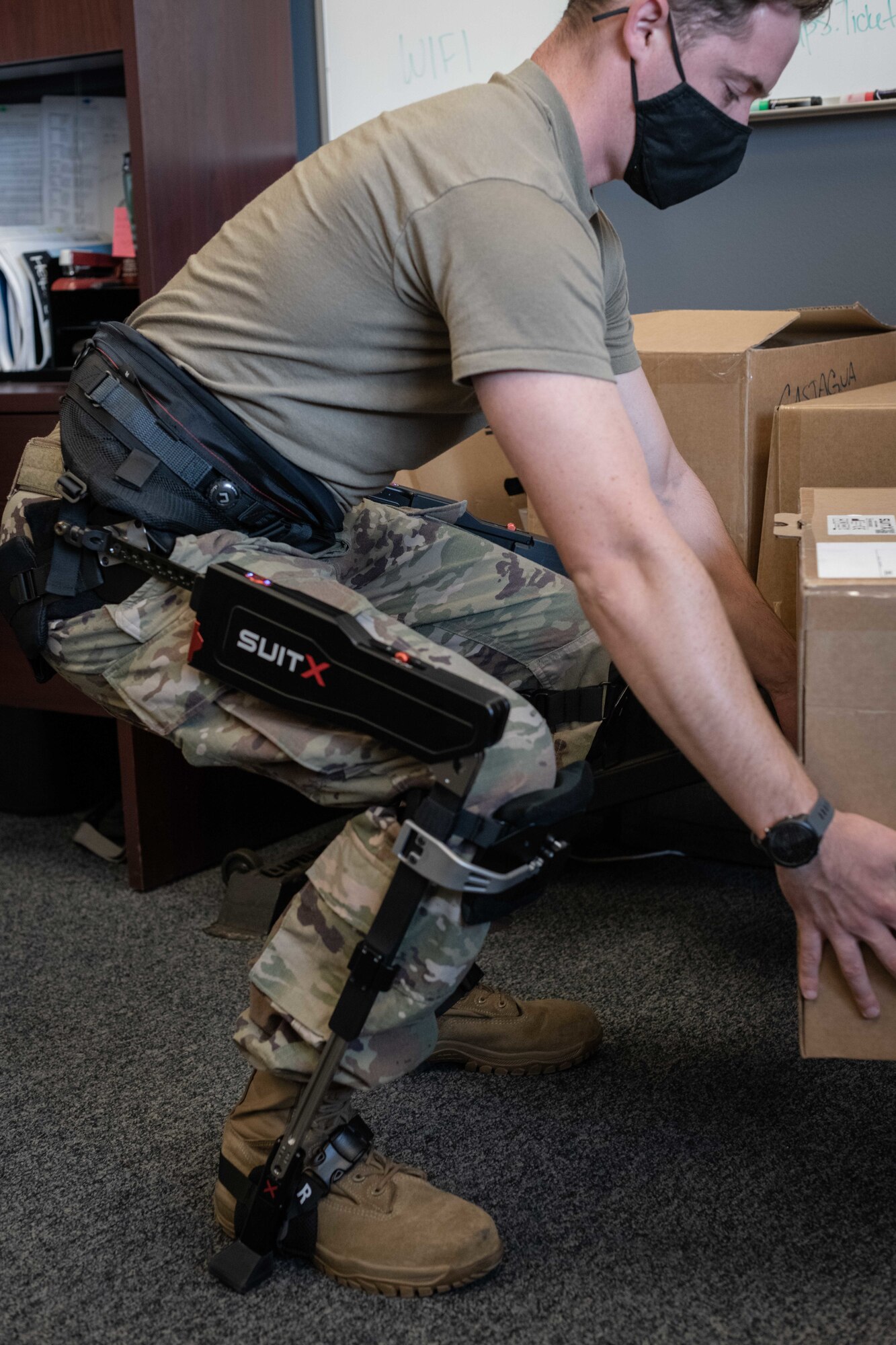 An Airman picks up a cardboard box while wearing a suit strapped to his hips, legs and feet.