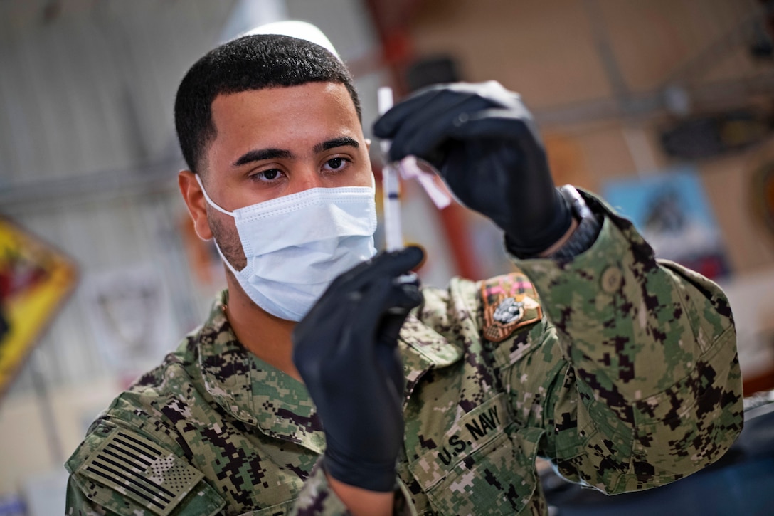 A male service member wearing a face mask and gloves looks at a syringe.